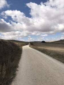 A gravel road leading off into the distance over low hills, with brown grassy fields on both sides, and a blue sky filled with fluffy clouds