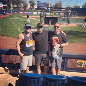 Matt Miller posing with two other men at a baseball game with the baseball field behind them
