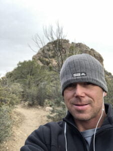 Selfie of Matt Miller with a rocky hill and a dirt path with vegetation behind him