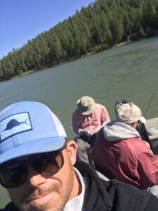 Selfie of Matt Miller on a boat with people sitting behind him, a lake, and distant tree-covered land in the background