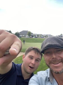 Selfie of Matt Miller with a young man pointing at the camera, holding a golf ball, with a golf course in the background