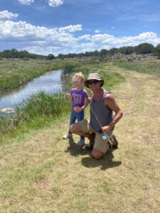 Matt Miller in a photo with a little girl holding a fishing rod near a green grass-covered land with a creek, trees in the distance, under a blue sky with white fluffy clouds