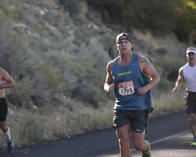 Matt Miller participating in a marathon, running on a road near a hill filled with desert shrubs and rocks, surrounded by other runners
