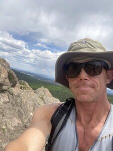 Selfie of Matt Miller in sunglasses and a hat on a rocky mountain, overlooking a green valley with trees under a cloud-filled blue sky